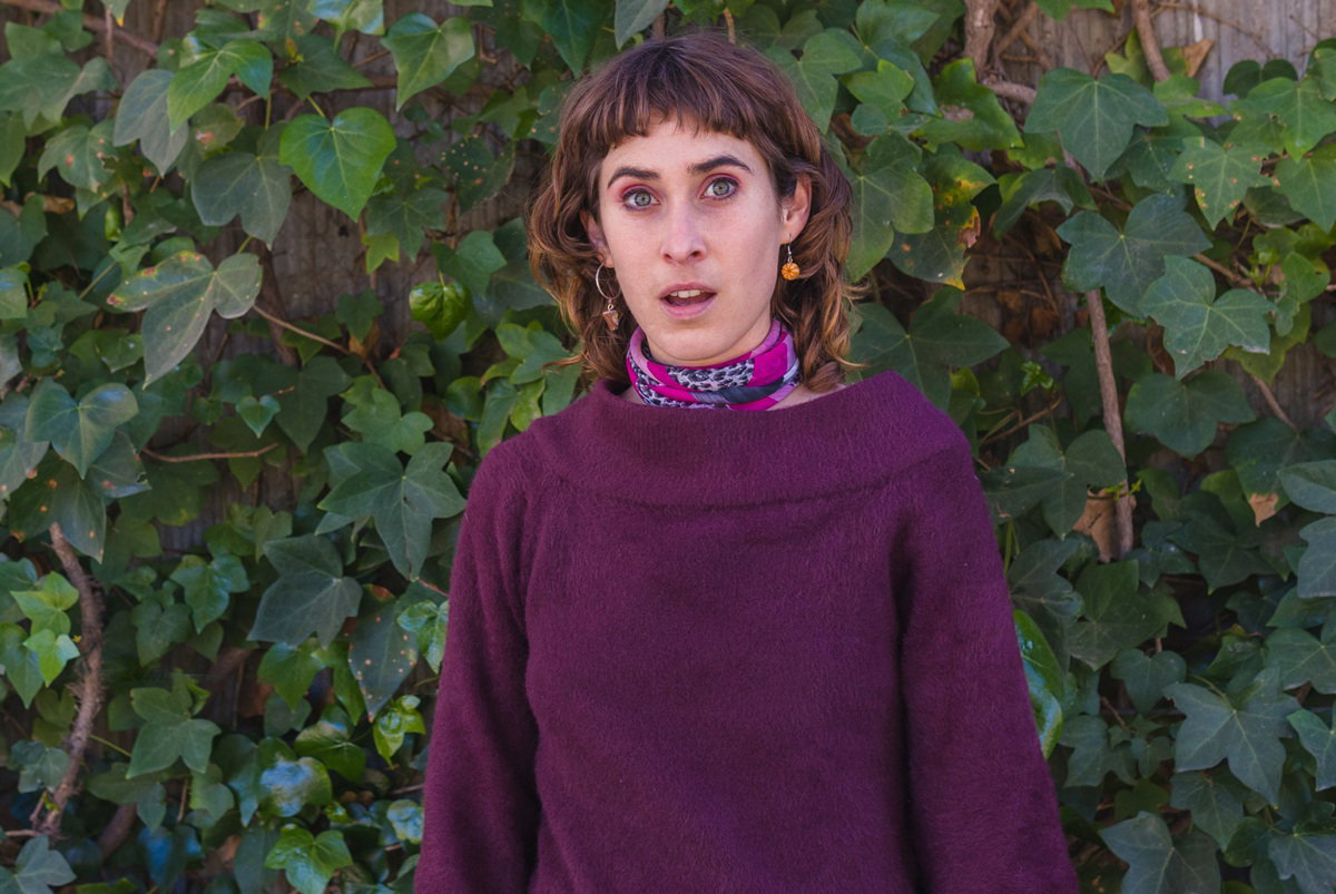 kobieta w wieku 30 plus w bakłażanowym swetrze z rozdziawioną buzią stoi przerażona na tle bluszczu