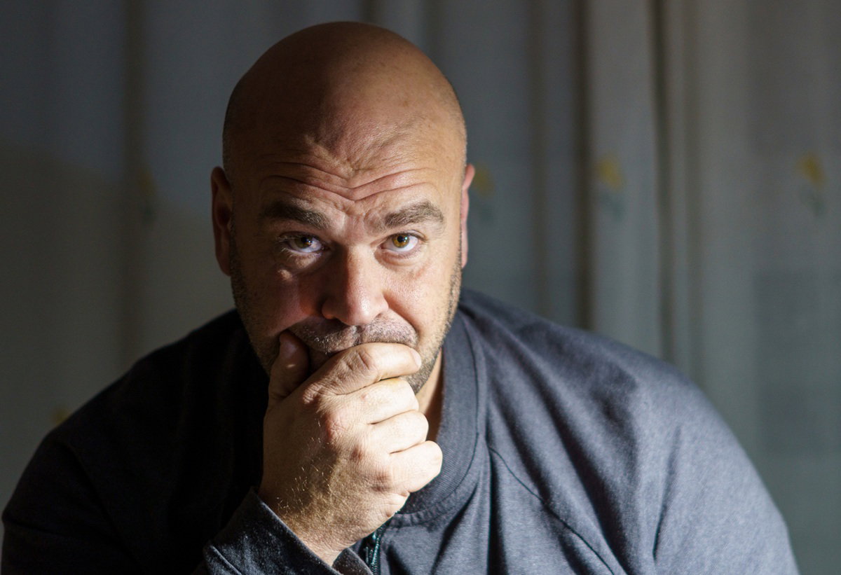 mężczyzna w wieku 35 plus łysy w bluzie siedzi wściekły w pokoju na tle zasłon