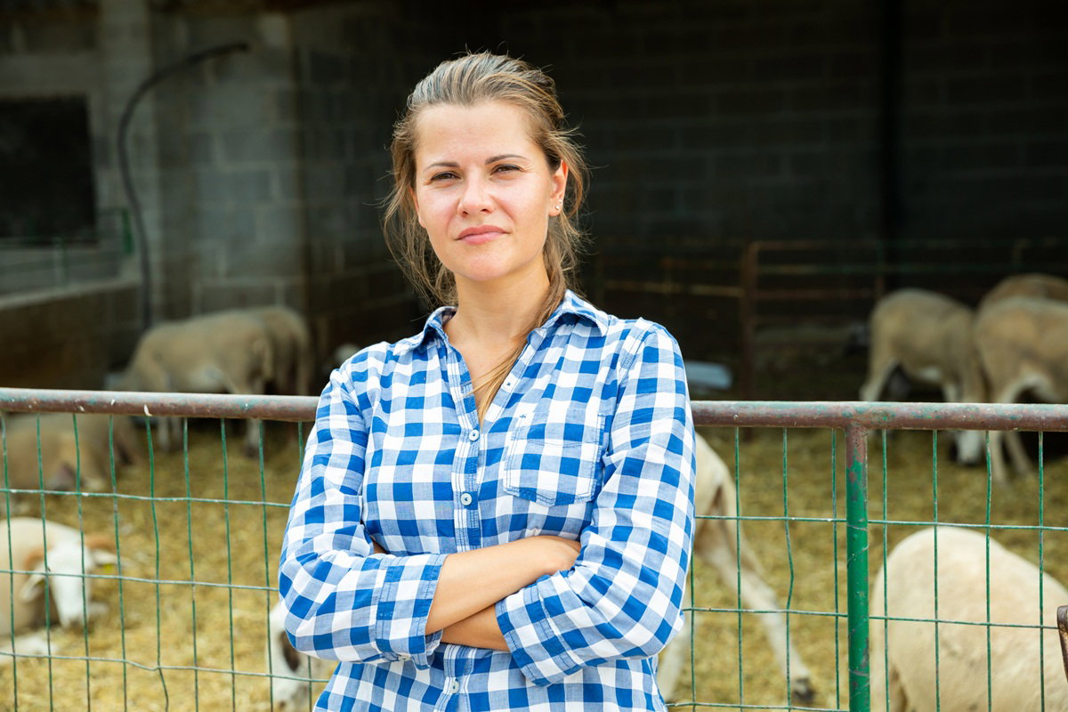 młoda kobieta w wieku 30 plus w kraciastej koszuli na tle zagrody ze zwierzętami stoi zgorzkniała