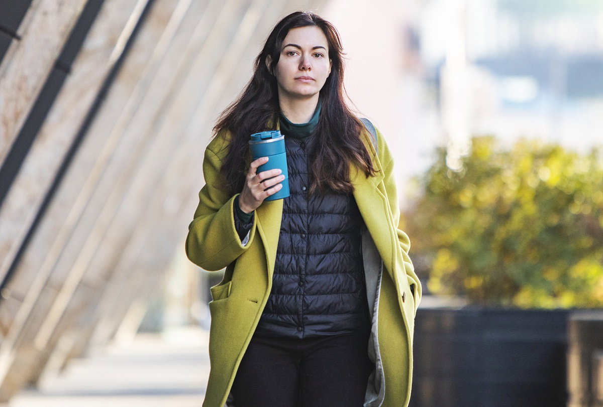 kobieta 35 plus z kubkiem kawy w ręku i w zielonym płaszczu spaceruje na zewnątrz zamyślona
