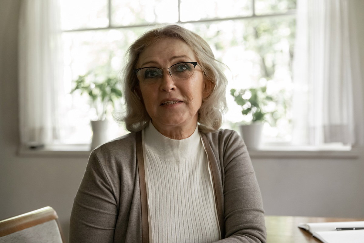 kobieta w wieku 60 plus w okularach siedzi przy stole na tle okna niepewna i zasmucona