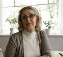 kobieta w wieku 60 plus w okularach siedzi przy stole na tle okna niepewna i zasmucona