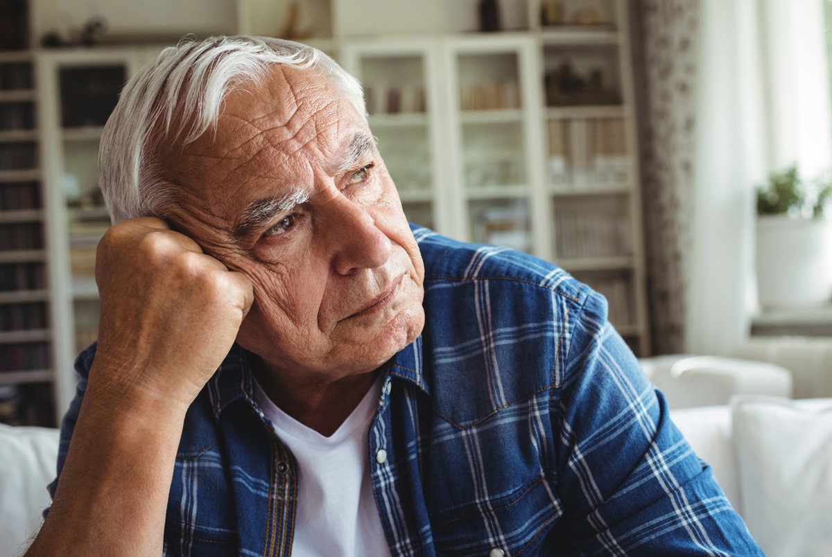 mężczyzna w wieku około 70 lat w niebieskiej koszuli w kratę siedzi w salonie smutny i zamyślony