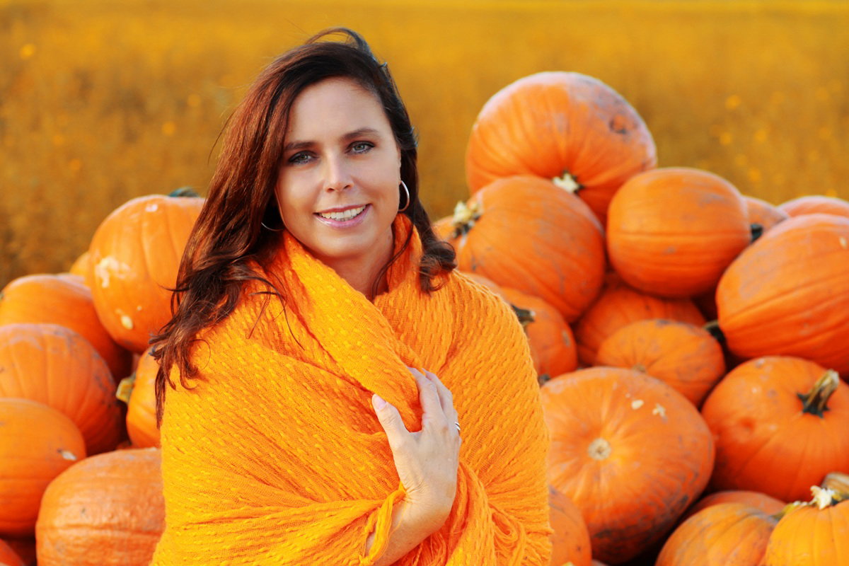 kobieta w wieku około 25 plus siedzi na tle dyń w pomarańczowym szalu i lekko się uśmiecha