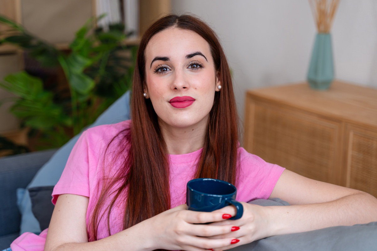 kobieta w wieku 25 plus w różowej koszulce siedzi na kanapie z kubkiem w ręku z dziwnym uśmiechem