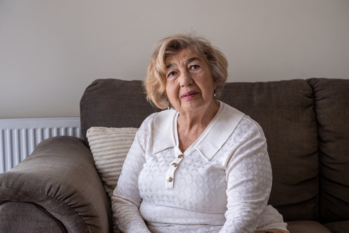kobieta w wieku około 70 lat w białej bluzce z kołnierzykiem siedzi na kanapie poważna i załamana