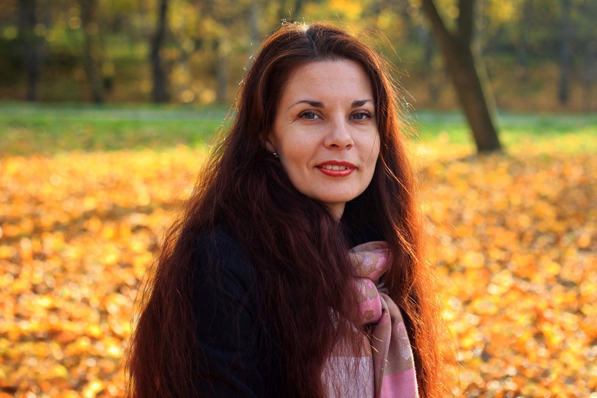 kobieta około 40 lat z długimi włosami siedzi na zewnątrz na tle jesiennego krajobrazu i lekko się uśmiecha