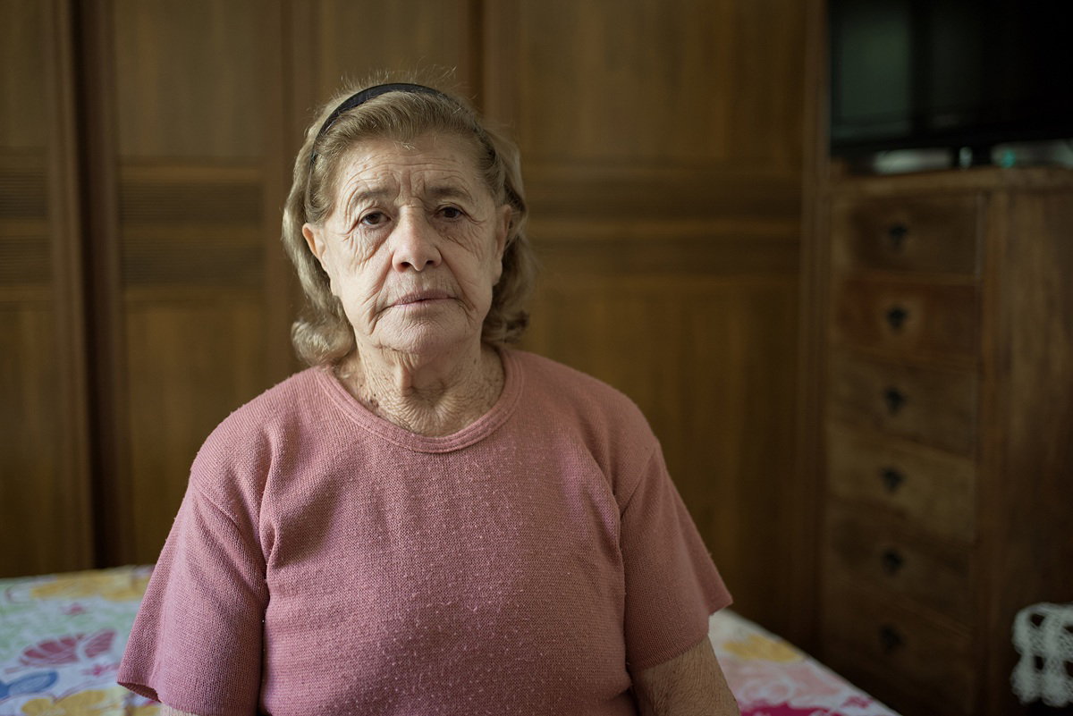 kobieta w wieku 70 plus siedzi smutna i zmartwiona na łóżku w pokoju na tle drewnianych szaf