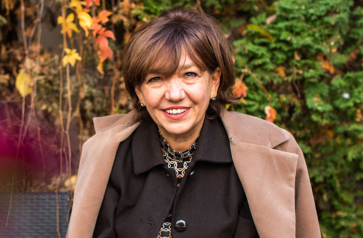 kobieta w wieku 60 plus siedzi w beżowym płaszczu na tle jesiennych liści i lekko się uśmiecha