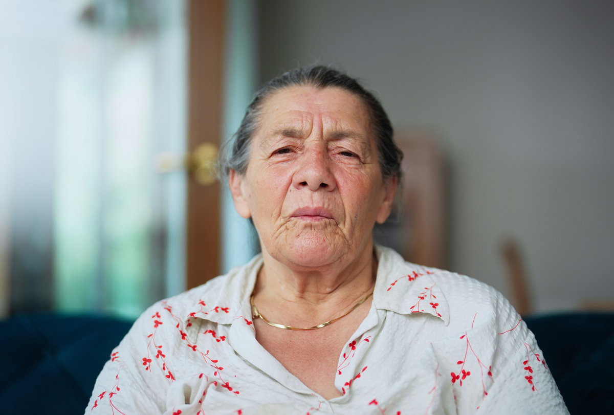 kobieta w wieku 70 plus w koszuli w kwiatowy wzór i z łańcuszkiem złotym na szyi siedzi z zaciętą miną zmartwiona
