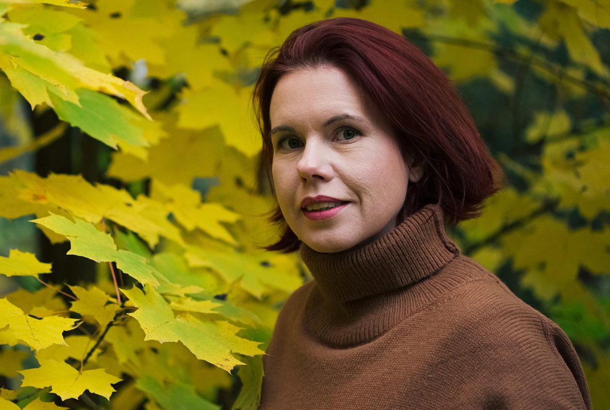 kobieta w wieku 35 plus w golfie karmelowym stoi lekko zaskoczona na tle żółtych liści