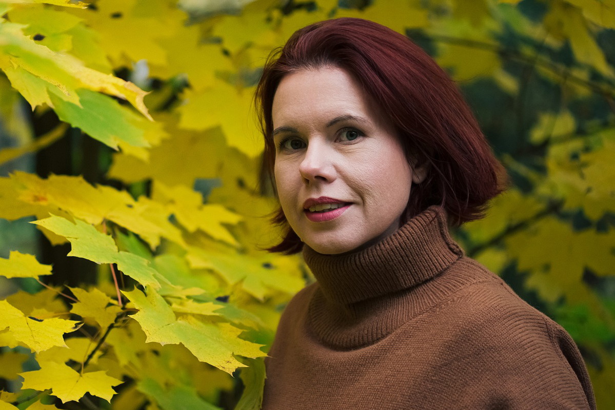 kobieta w wieku 35 plus w golfie karmelowym stoi lekko zaskoczona na tle żółtych liści
