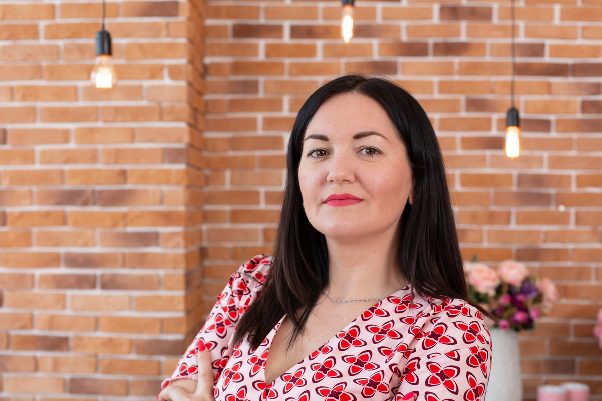 kobieta w wieku około 35+ z czarnymi włosami i we wzorzystej sukience stoi na tle ceglanej ściany z poważną miną