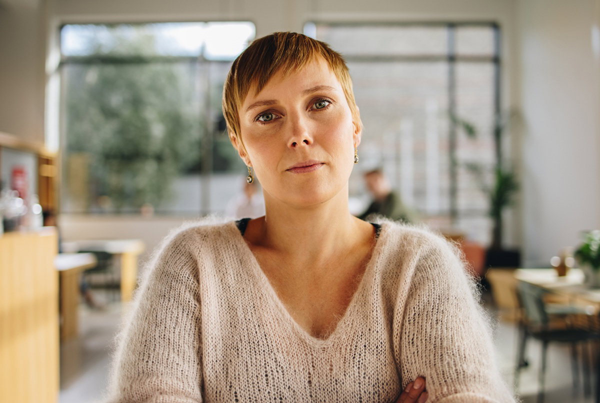 kobieta w wieku około 30 lat w sweterku z dekoltem i z krótkimi włosami stoi smutna i poważna na tle biura