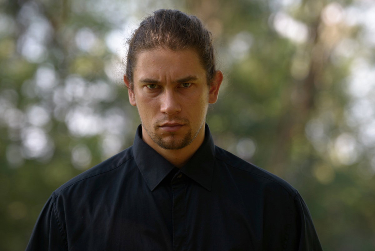 mężczyzna w wieku 30 plus w czarnej koszuli z długimi włosami zaczesanym do tyłu i  z zarostem stoina zewnaątrz zagniewany