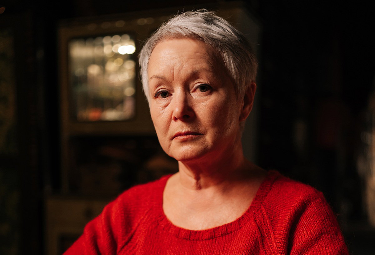 kobieta w wieku 55 plus krótkie siwe włosy w czerwonym swetrze siedzi w ciemnym pokoju ze smutną miną