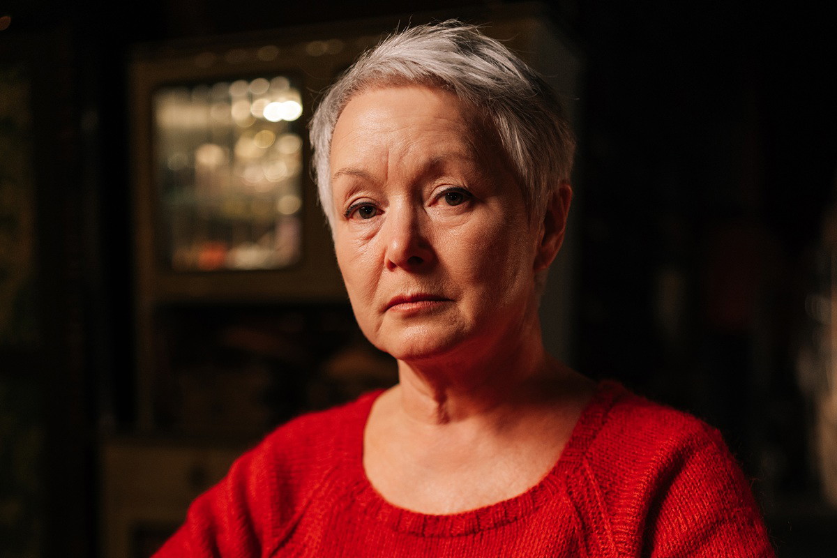 kobieta w wieku 55 plus krótkie siwe włosy w czerwonym swetrze siedzi w ciemnym pokoju ze smutną miną