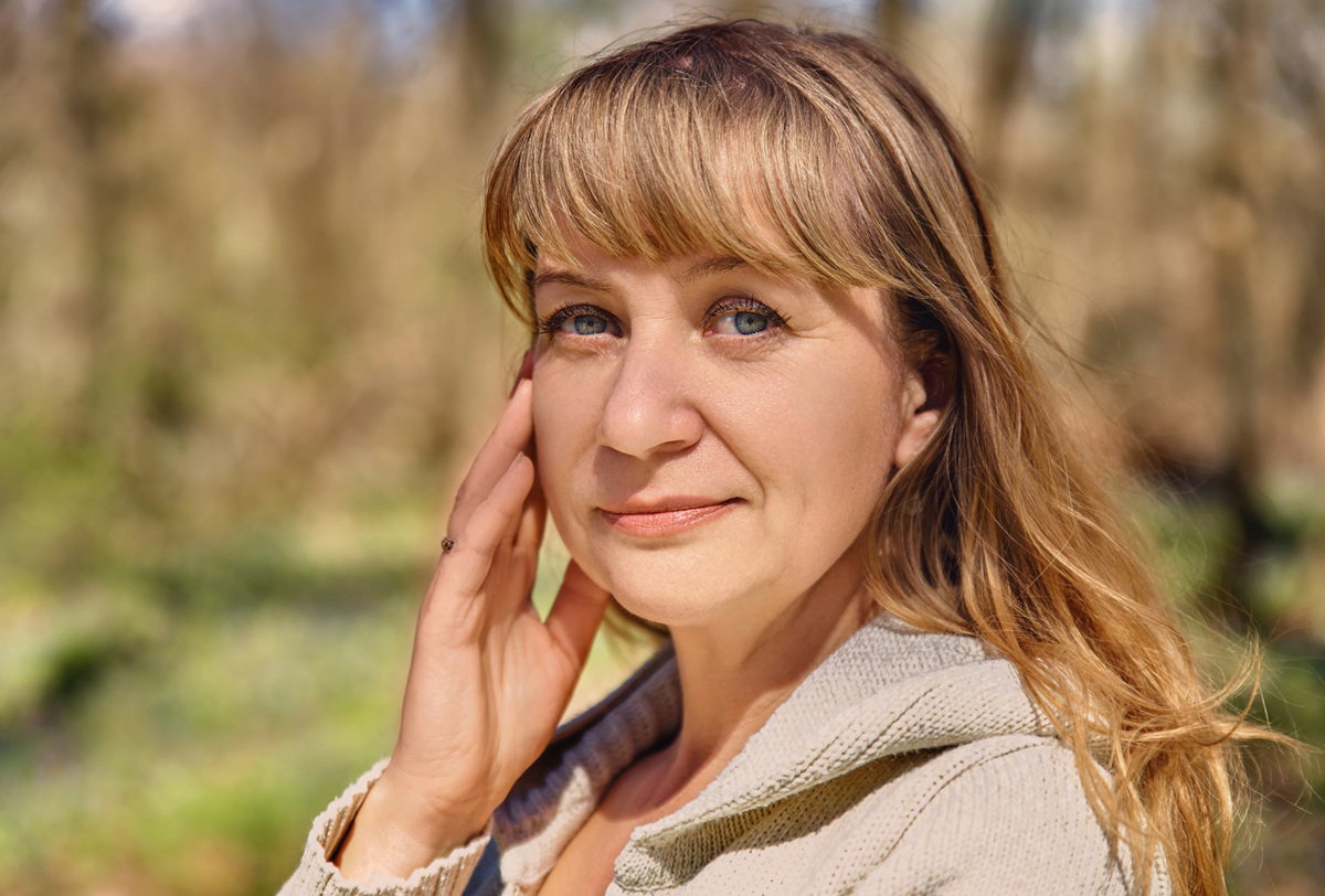 kobieta 45 plus blondynka w beżowym sweterku z grzywką stoi na zewnątrz zamyślona