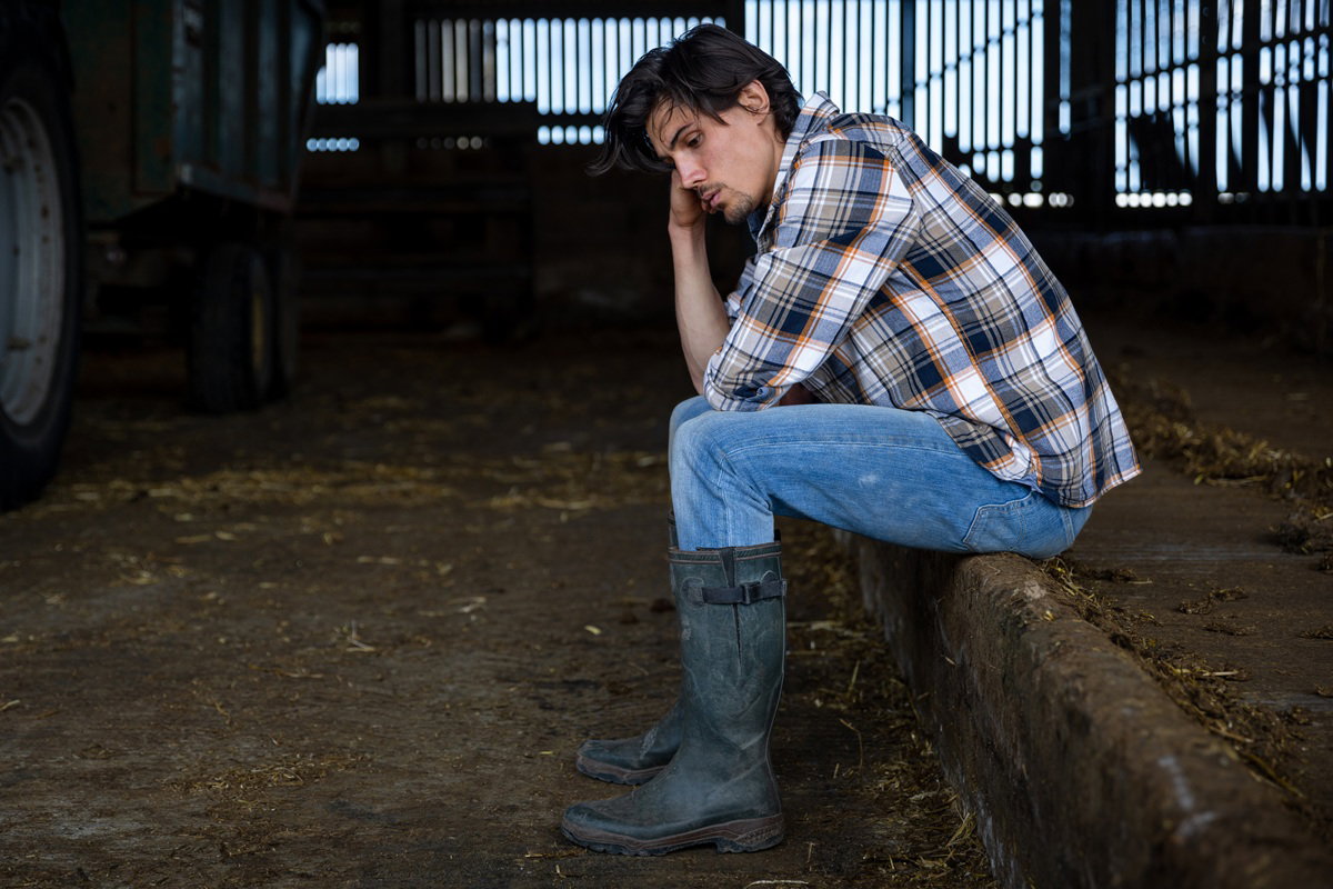 mężczyzna w wieku 25 plus siedzi przy traktorze wewnątrz w gumiakach smutny i rozmyśla