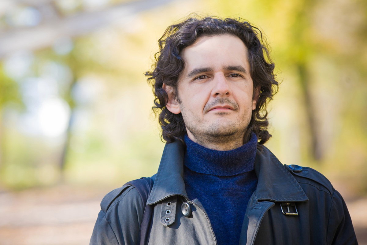 mężczyzna w wieku 35 plus z dłuższymi ciemnymi włosami i w kurtce stoi na zewnątrz zamyślony