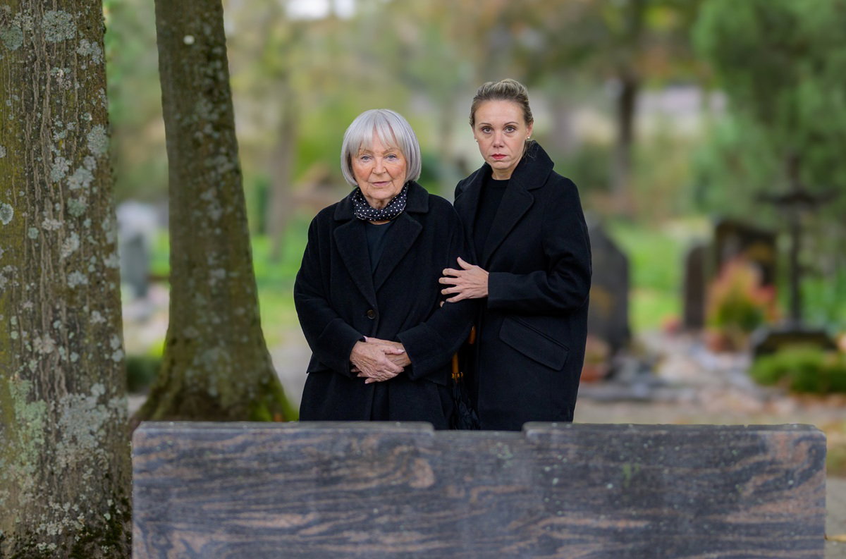 dwie kobiety stoją przy grobie zmarłego w czarnych strojach zamyślone i smutne