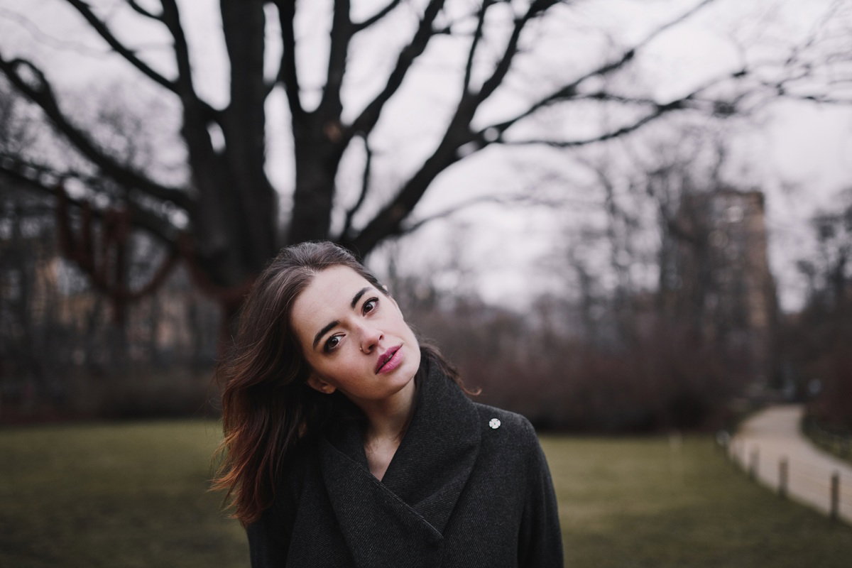 kobieta w wieku około 30 lat w płaszczu stoi na tle drzewa bez liści zamyślona