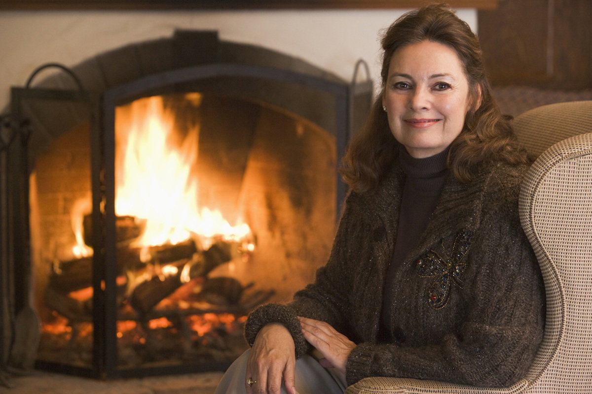 kobieta w wieku 60 plus w ozdobnym swetrze siedzi przy kominku na fotelu i uśmiecha się zadowolona