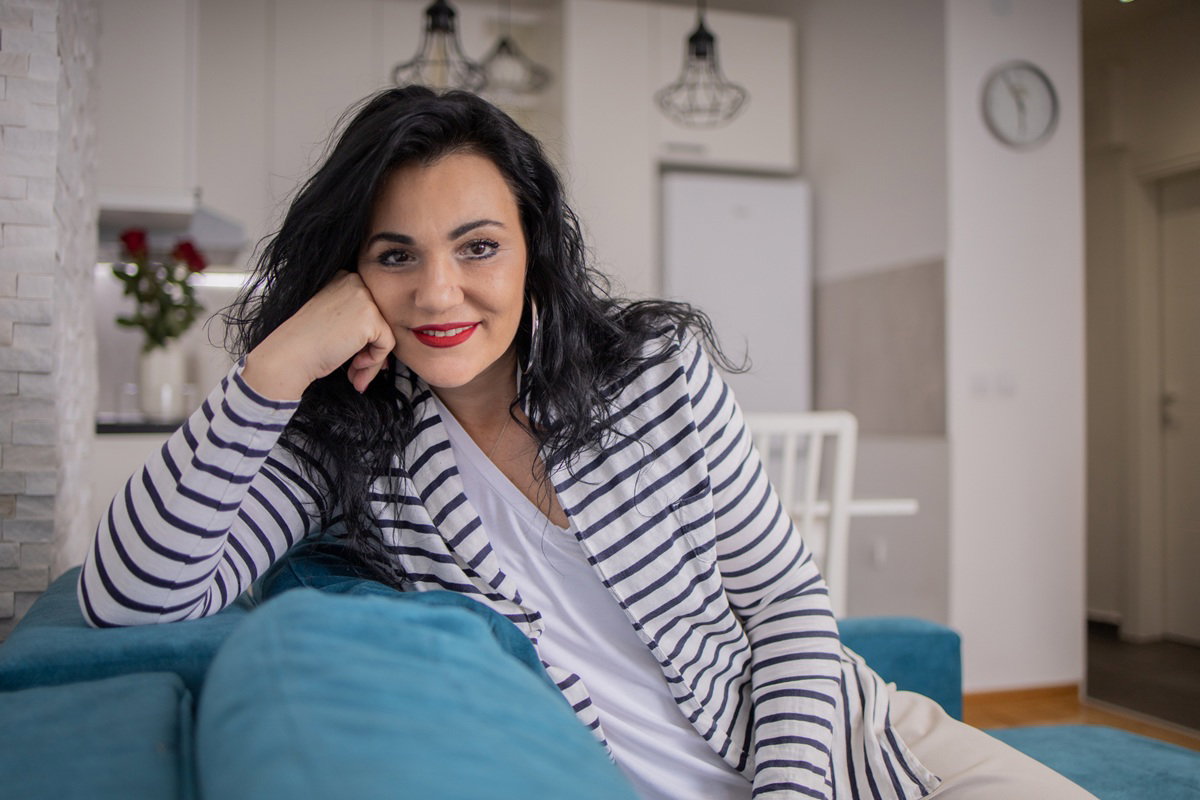 kobieta w wieku około 35 lat w pasiastym wdzianku i z ciemnymi włosami siedzi na kanapie i uśmiecha się rozmarzona