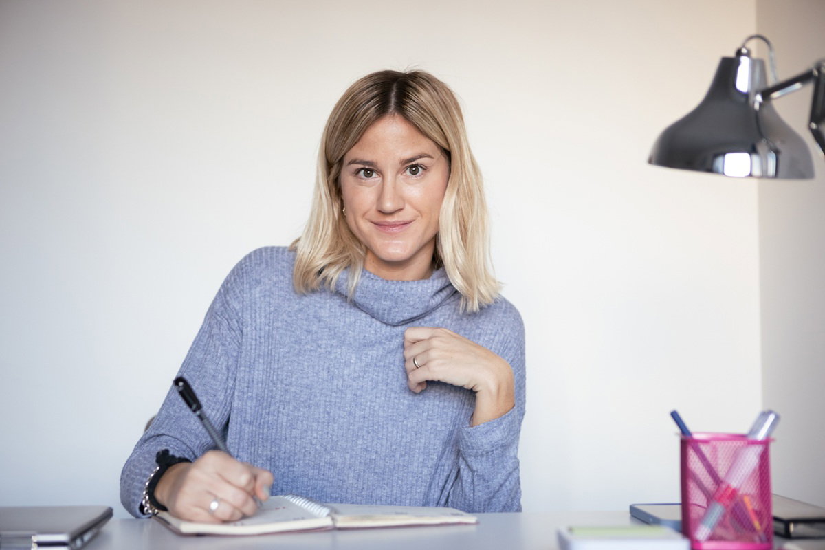 kobieta w wieku 30 plus w szarym swetrze siedzi przy biurku niepewna ale lekko się uśmiecha