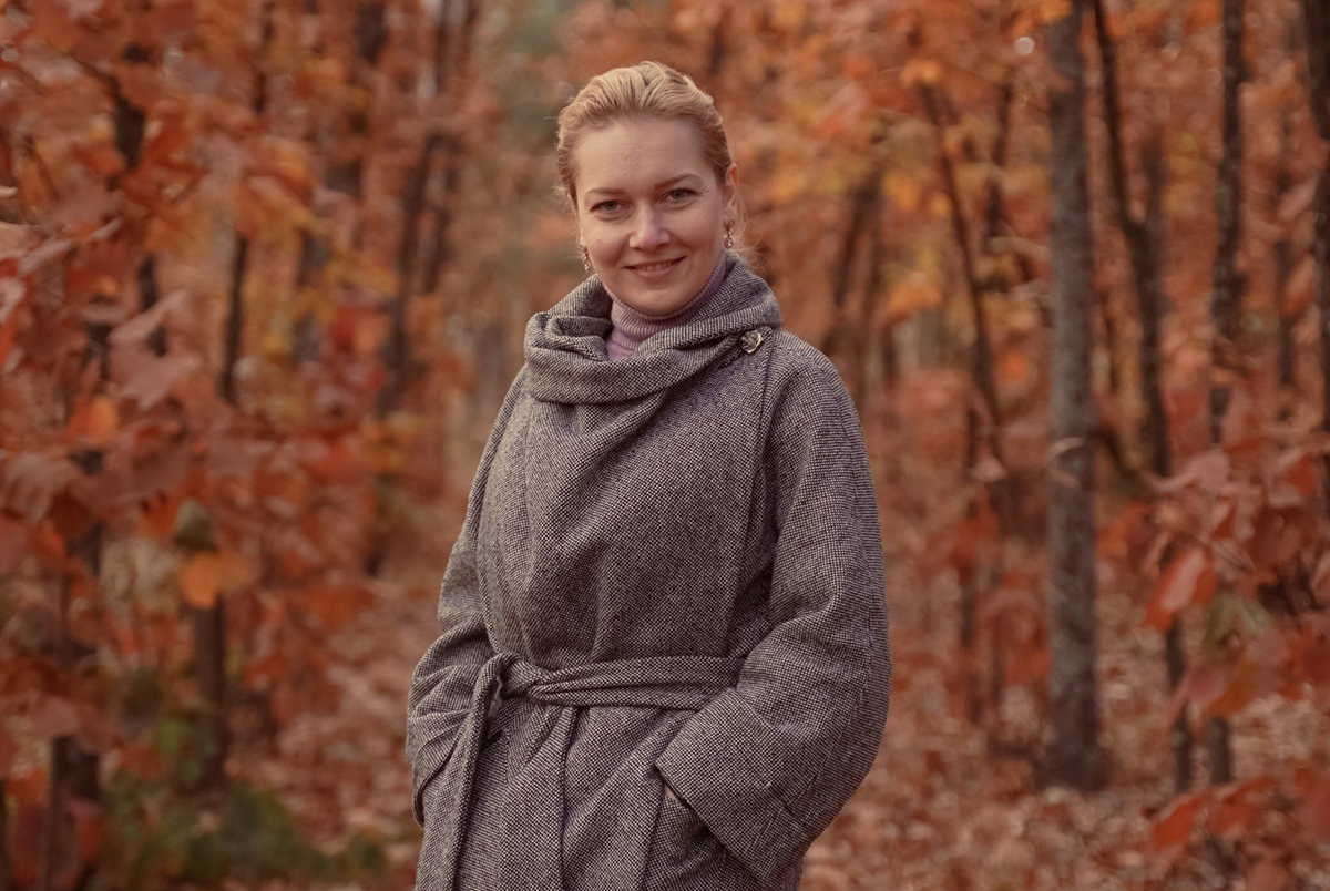 Kobieta w wieku 35 plus w szarym płaszczu na tle rudych liści z uśmiechem