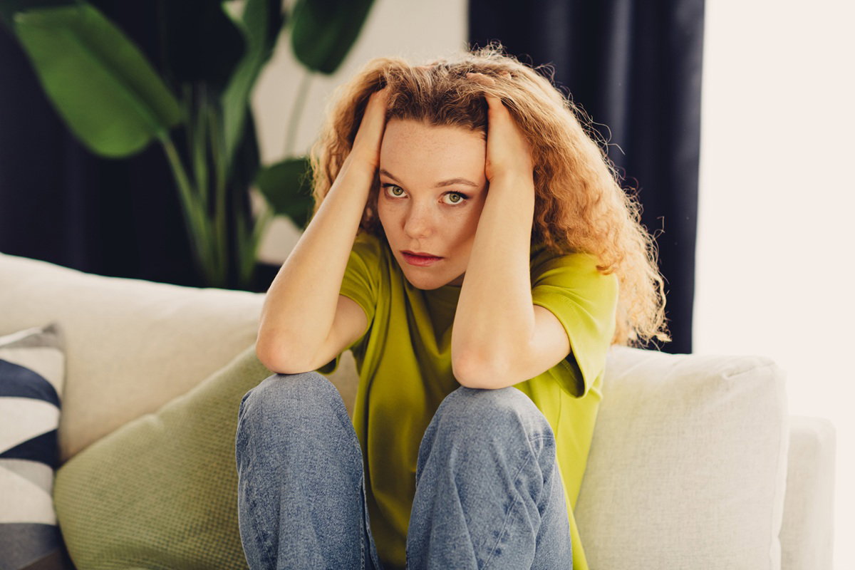 młoda kobieta w wieku 20 plus z długimi rudawymi kręconymi włosami siedzi na kanapie załamana