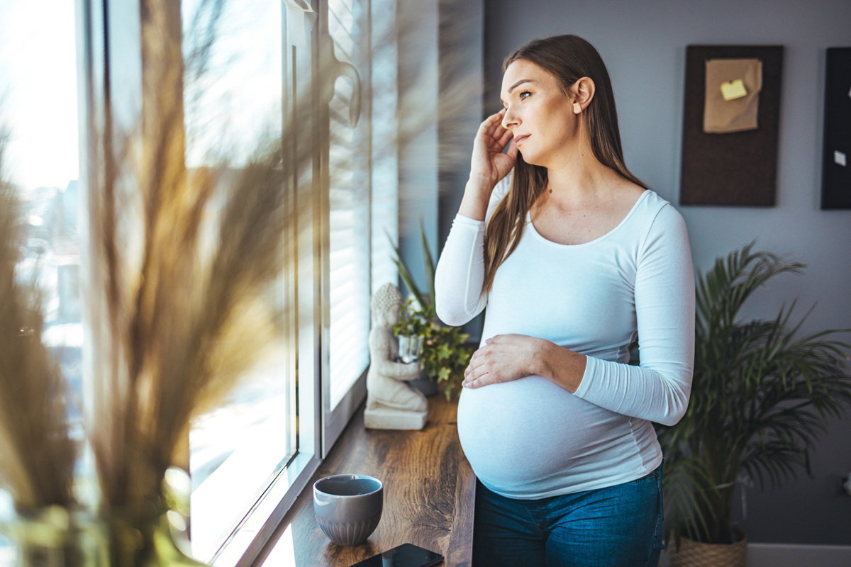 kobieta w wieku 30 plus w ciąży stoi zamyślona i smutna przy oknie