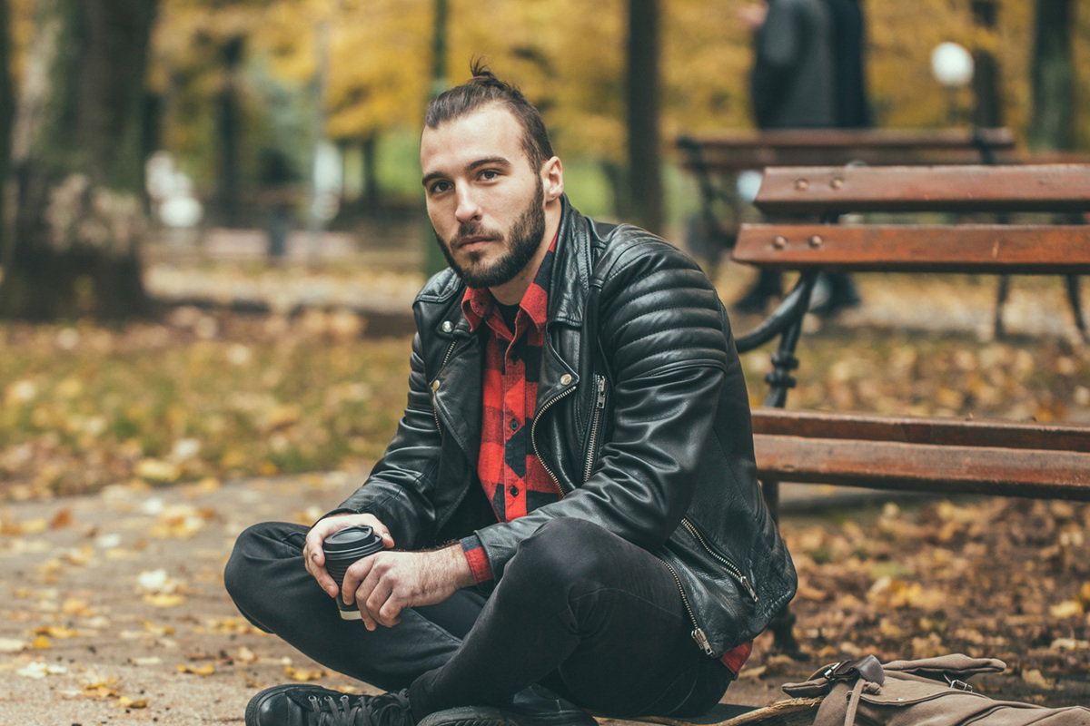 młody mężczyzna w wieku około 30 lat w skórzanej kurtce siedzi w jesiennym parku z kubkiem kawy w ręku