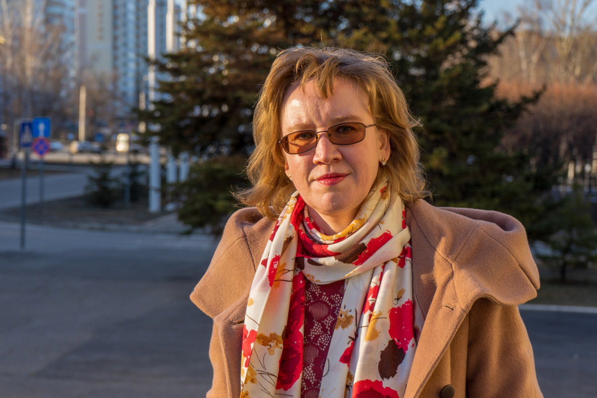 kobieta w wieku 55 plus w okularach i beżowym płaszczu stoi na tle bloków zdeterminowana i dumna z siebie