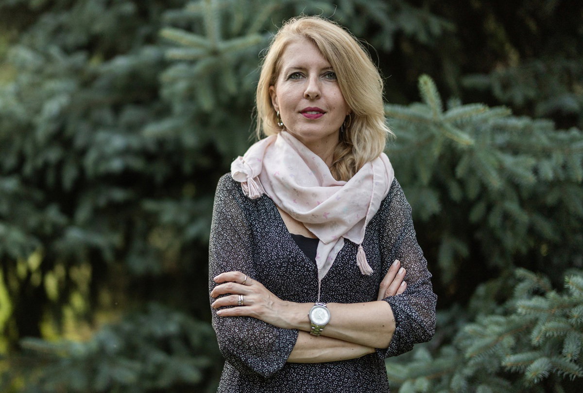 kobieta w wieku 45 plus blondynka stoi na tle drzew iglastych lekko podminowana