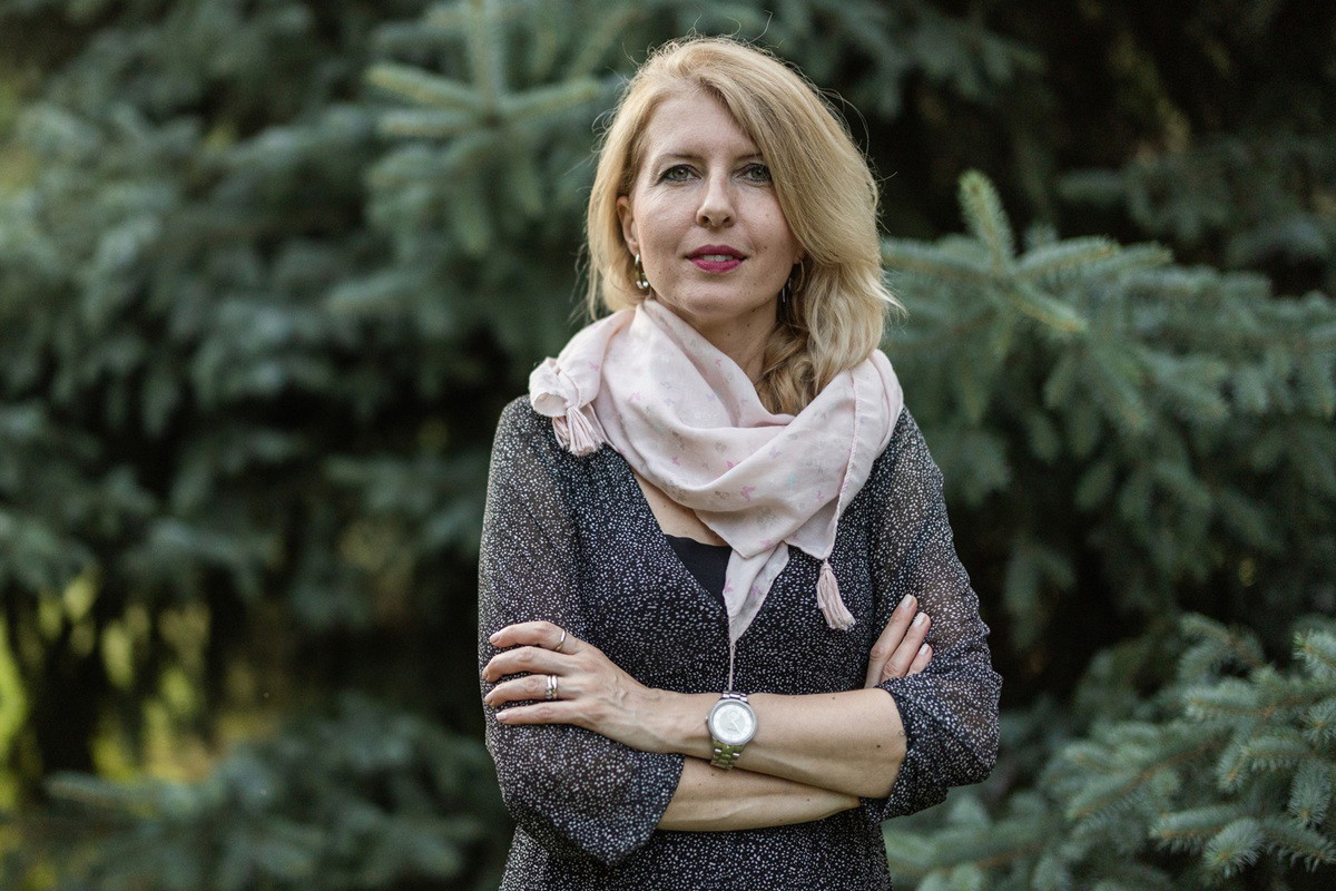 kobieta w wieku 45 plus blondynka stoi na tle drzew iglastych lekko podminowana