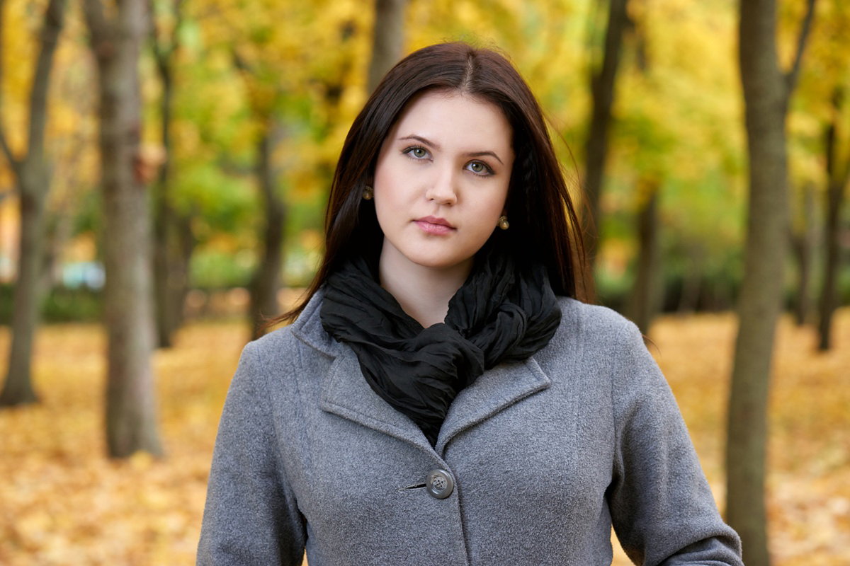 kobieta w wieku 20 plus z ciemnymi włosami i w szarym płaszczu stoi na tle jesiennego parku zamyślona i smutna