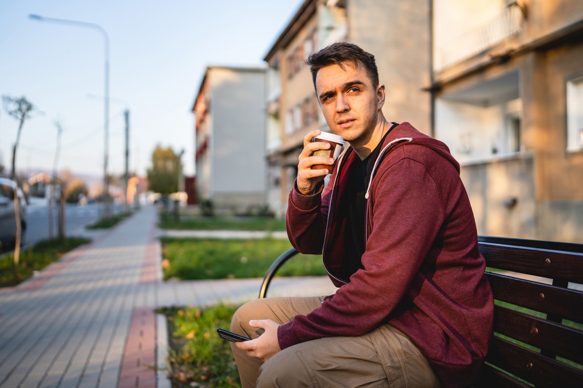 mężczyzna w wieku 25 plus w bluzie z kapturem siedzi na ławce z telefonem w dłoni i pije kawę w kubku