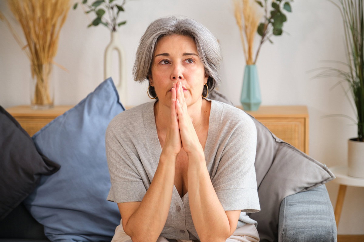 kobieta w wieku około 50 lat z siwymi włosami i ze złotymi kolczykami siedzi an kanapie i się zamartwia