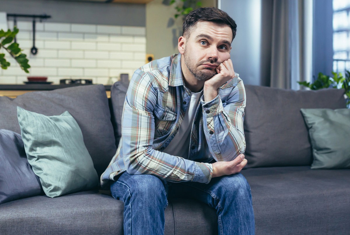 mężczyzna w wieku 30 plus w kraciastej koszuli siedzi zmartwiony na kanapie na tle kuchni