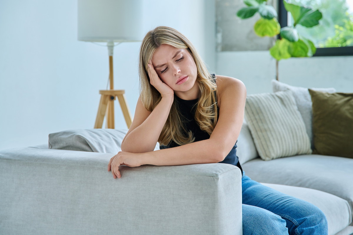 Unhappy upset young woman having headache, sitting on sofa at home