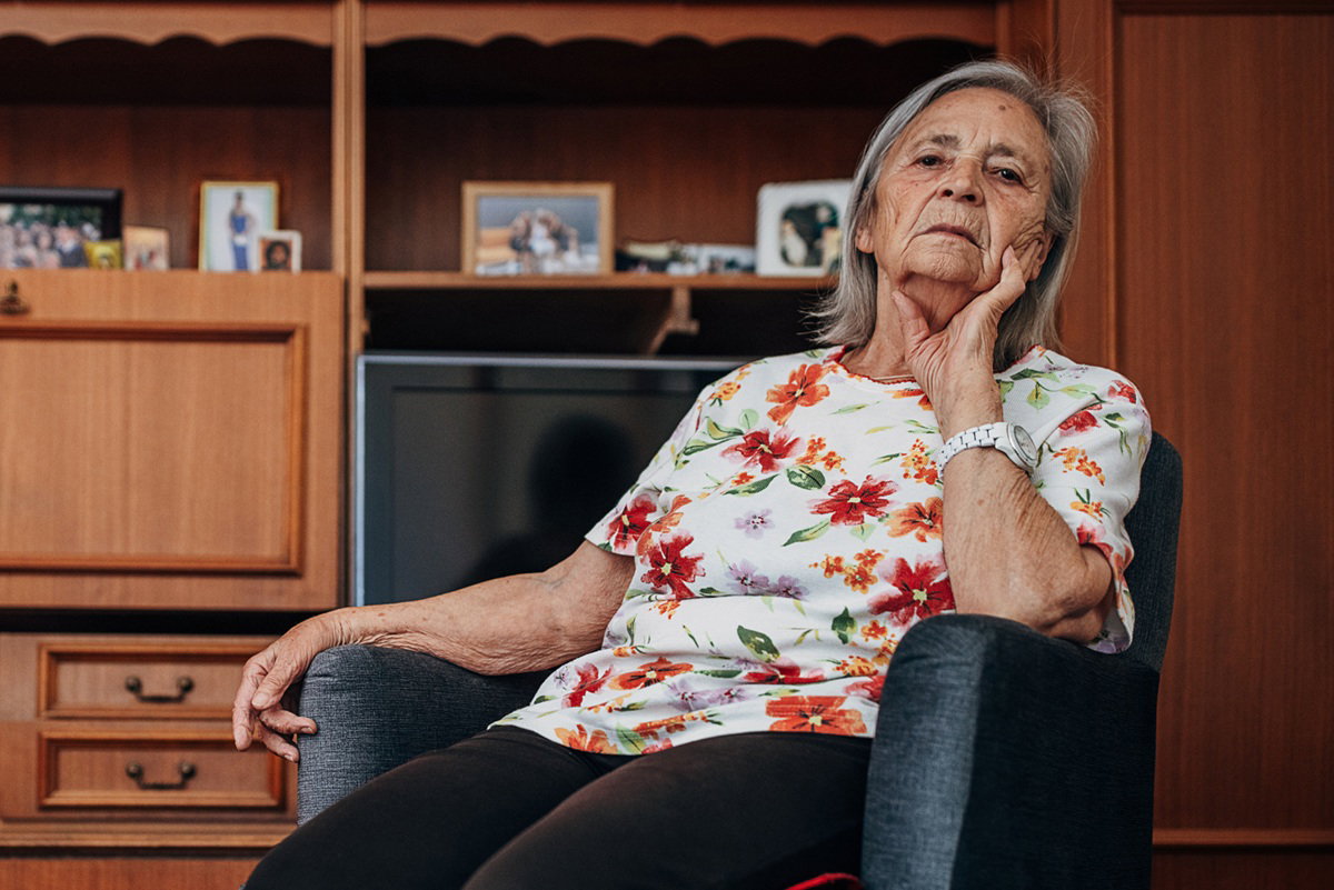 kobieta w wieku 70 plus siedzi na fotelu na tle meblościanki i telewizora zamyślona i poważna