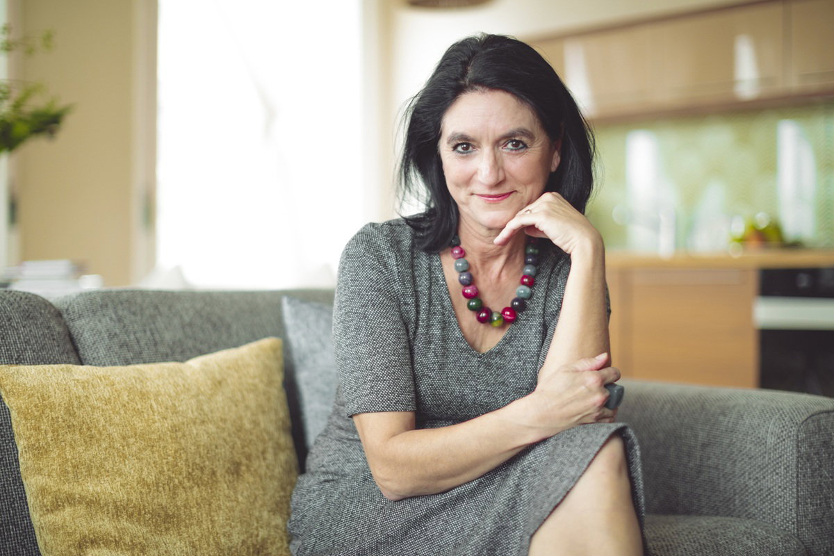 kobieta w wieku 50 plus z kolorowymi koralami na szyi siedzi na kanapie i uśmiecha się zalotnie