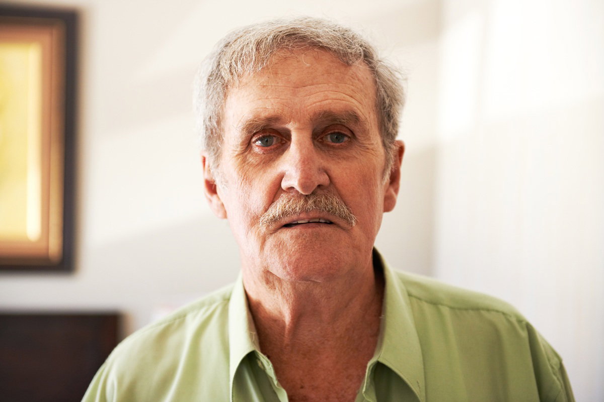 mężczyzna w wieku około 70 lat z wąsem i w zielonej koszuli stoi smutny i zgorzkniały