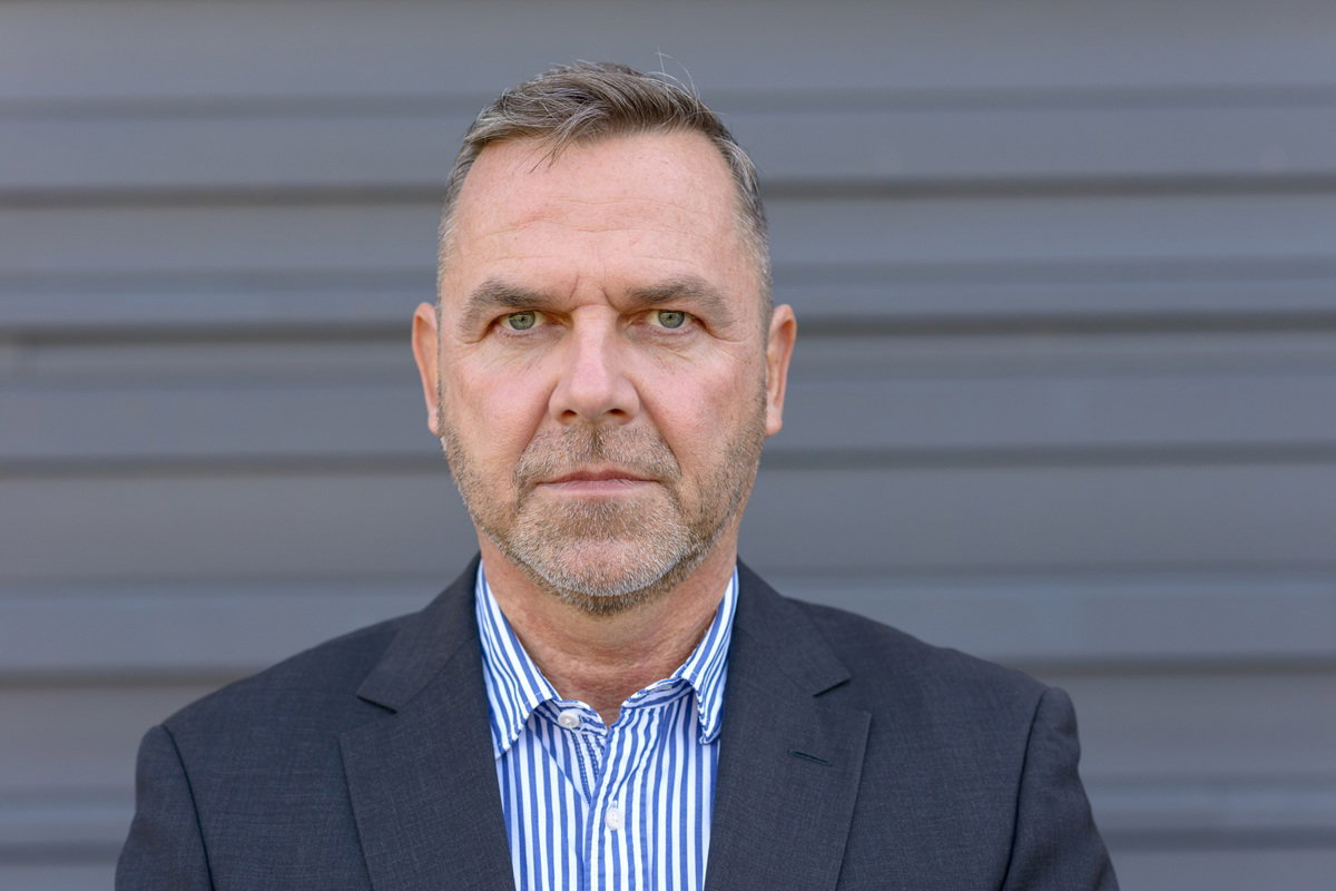 mężczyzna w wieku około 40 lat w pasiastej koszuli i ciemnej marynarce stoi zamyślony i poważny na tle metalowej szarej ściany