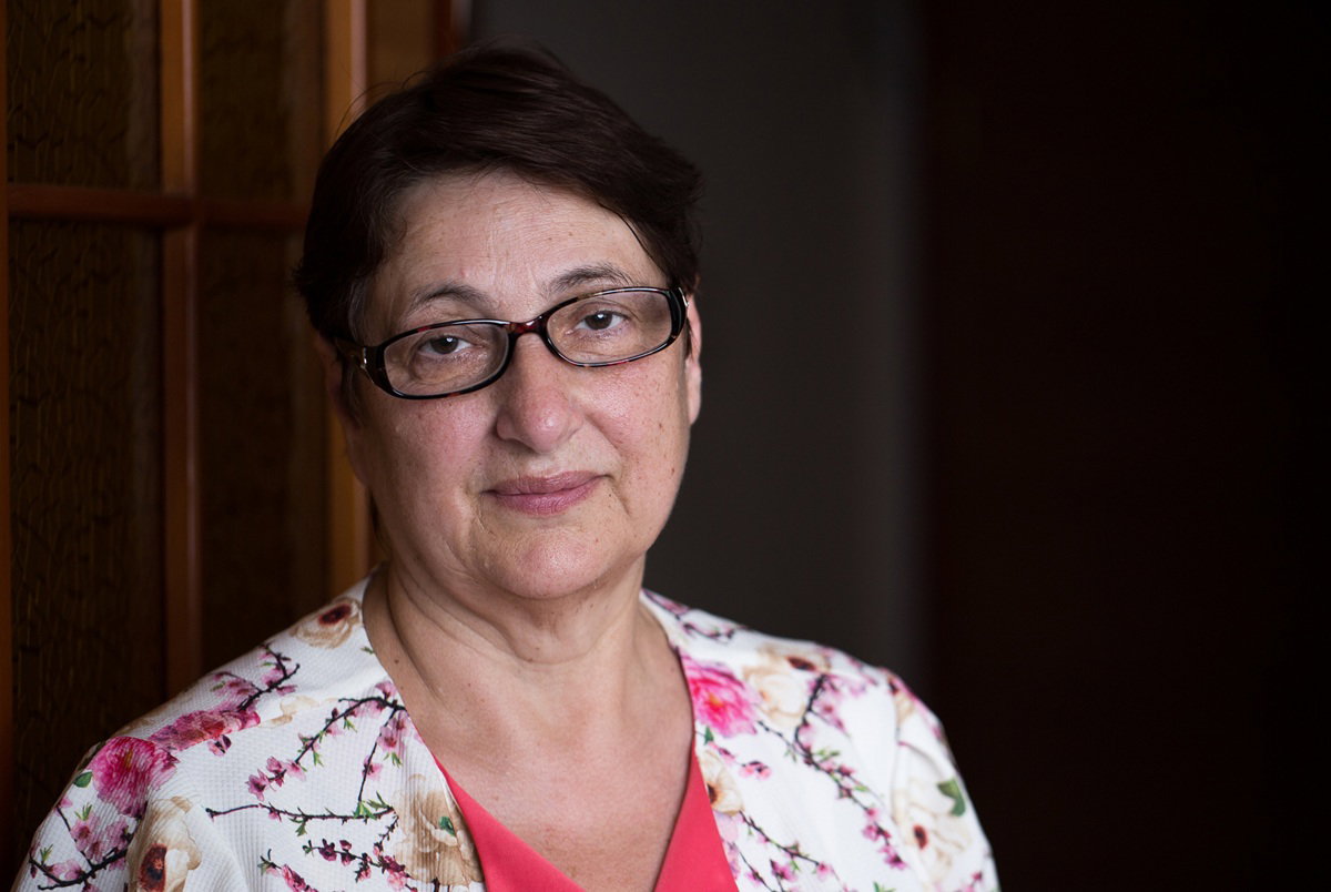 kobieta w wieku 50 plus w kwiecistej narzutce i w okularach stoi na tle drzwi i patrzy znużonym wzrokiem