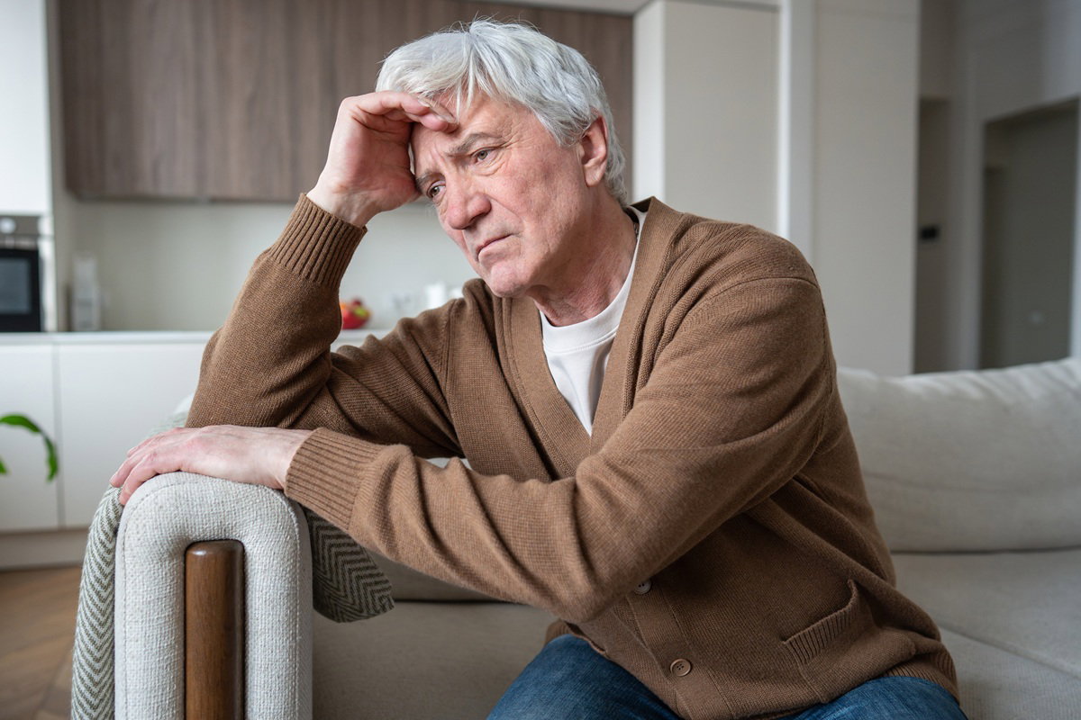 mężczyzna w wieku 60 plus w karmelowym swetrze siedzi zamyślony i smutny na kanapie