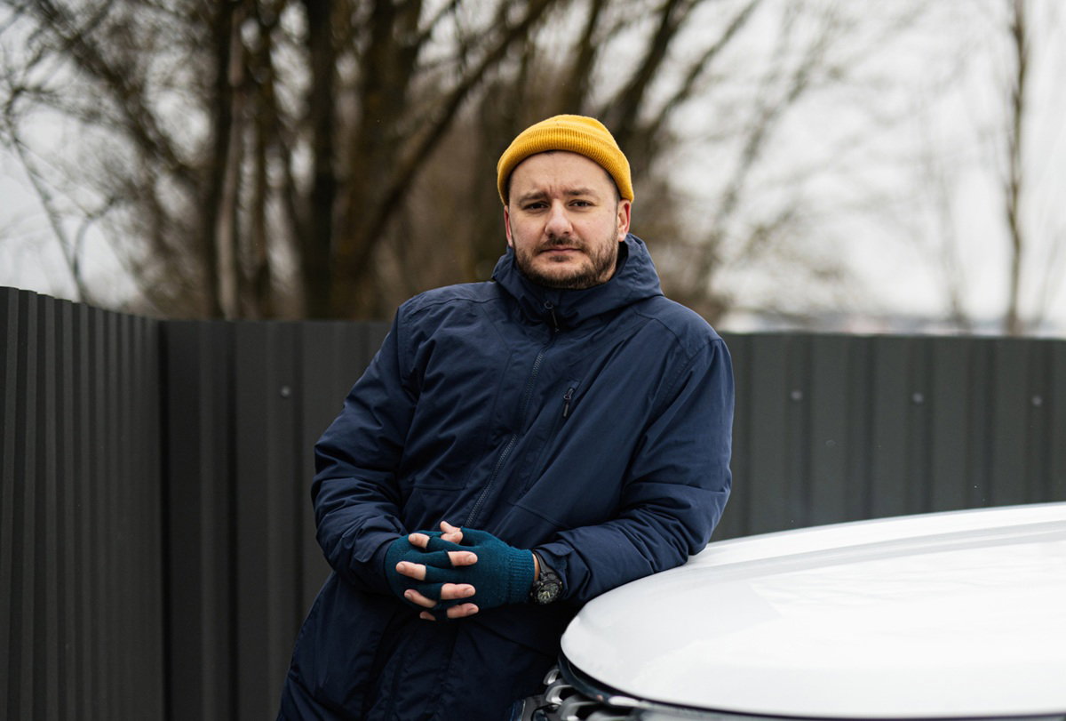 mężczyzna w wieku 35 plus w żółtej czapce z zarostem w granatowej kurtce stoi na zewnątrz zmartwiony i opiera się o samochód