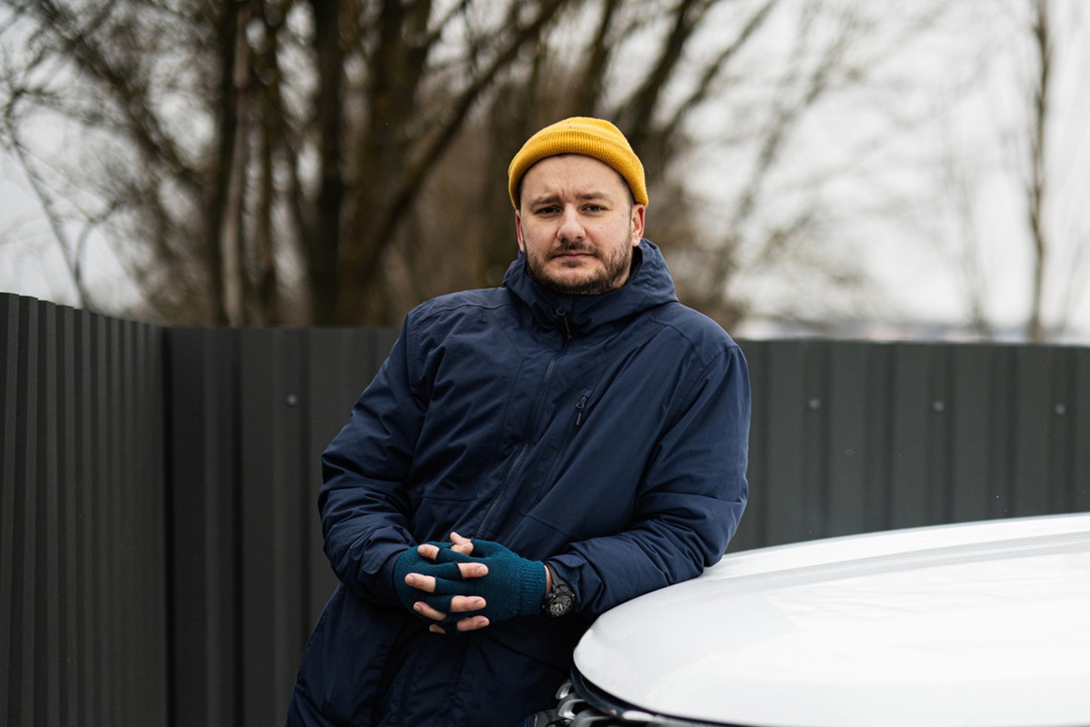 mężczyzna w wieku 35 plus w żółtej czapce z zarostem w granatowej kurtce stoi na zewnątrz zmartwiony i opiera się o samochód