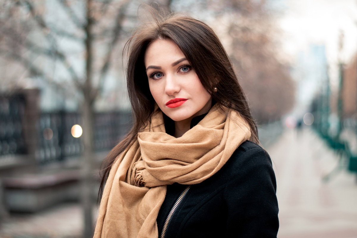 kobieta w  wieku 25 plus w beżowym szalu i z czerwoną szminką stoi zamyślona na ulicy
